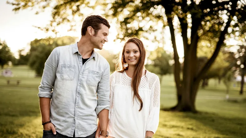 Young couple holding hands and smiling in a sunlit park with trees and grass in the background