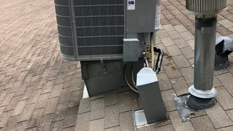 Gray rooftop HVAC unit installed on brown shingle roof with nearby vent pipes and green trees in background.
