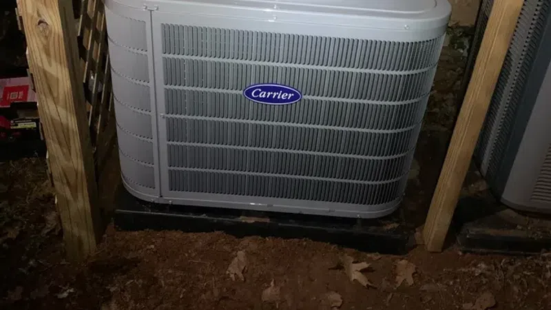Carrier air conditioning unit installed outdoors beneath wooden framing on dirt ground at night.