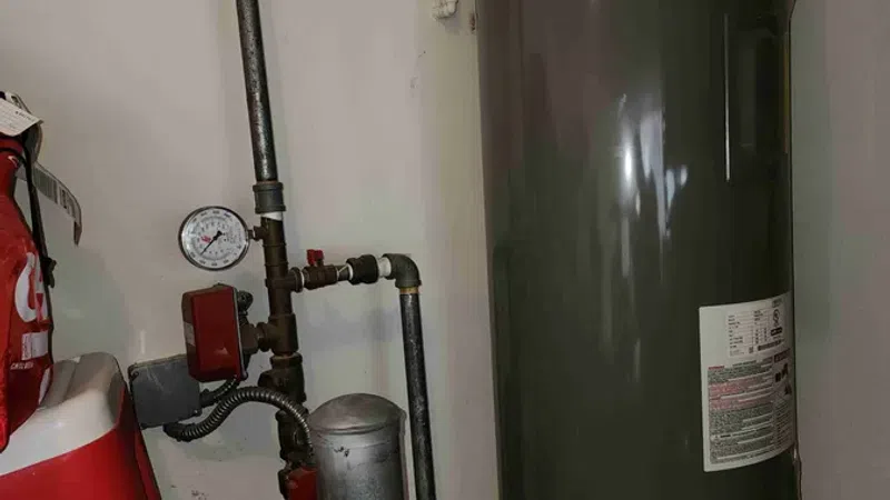 Tall gray water heater tank with pipes and pressure gauge installed in a utility room corner.