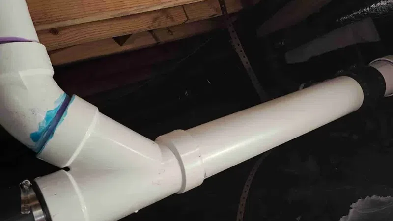 White PVC plumbing pipes connected in a crawl space under wooden floor joists with insulation visible.