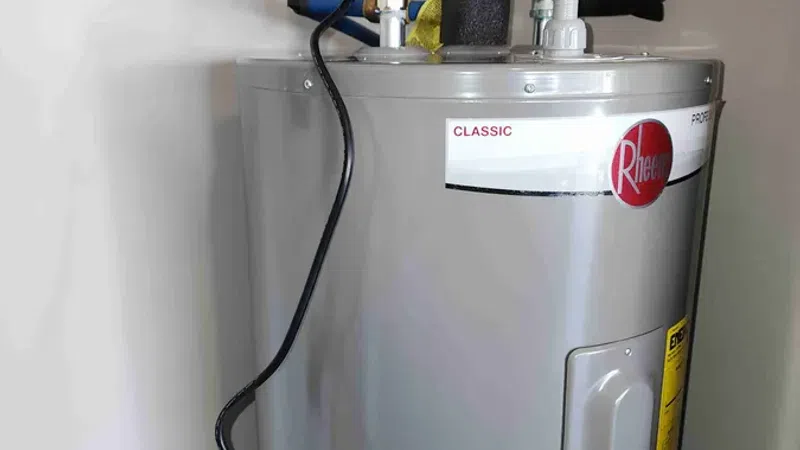 Gray Rheem Classic water heater with attached red circulation pump installed in a corner of the room.