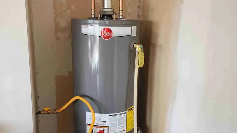 Gray Rheem water heater with connected copper and yellow pipes in a utility room corner.