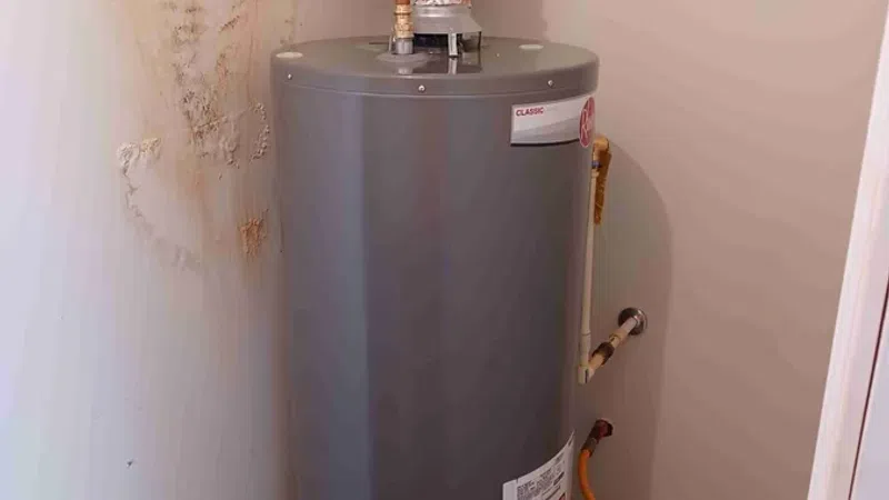 Gray water heater with copper pipes installed in a small utility closet with wall damage nearby.