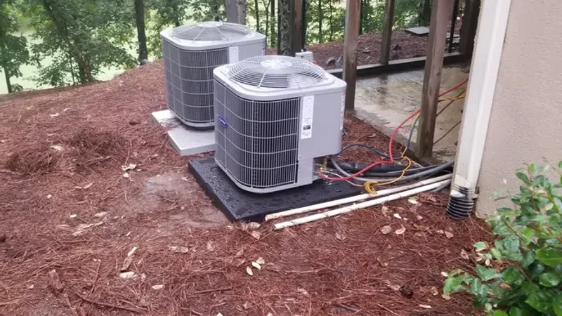 Two outdoor air conditioning units installed on concrete pads beside a house with mulch ground and surrounding trees.