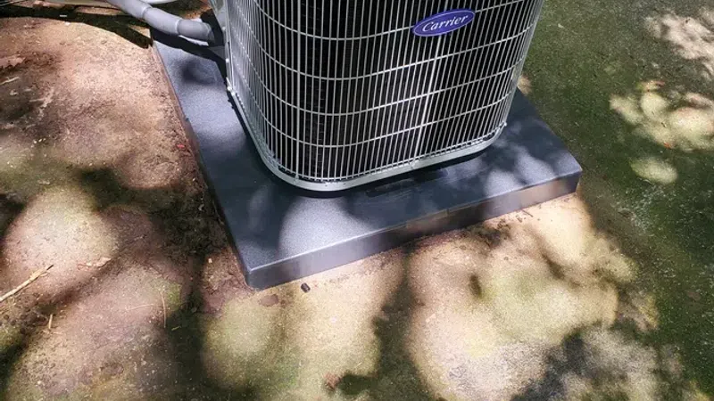 Outdoor Carrier brand air conditioning unit installed on black platform beside house wall with shaded ground.
