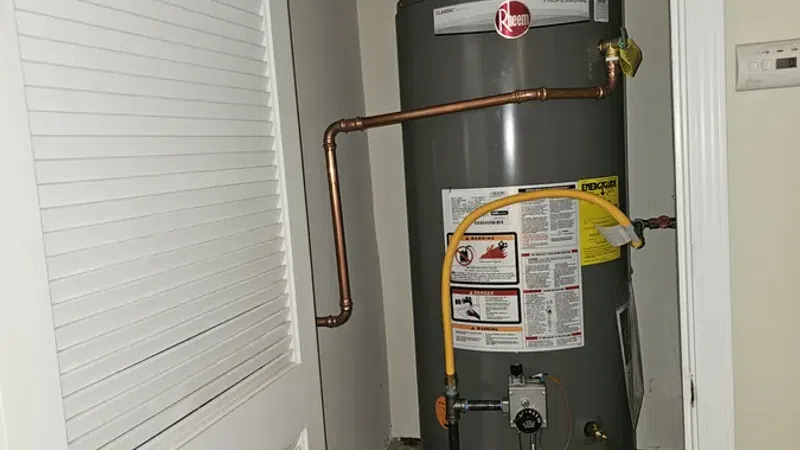 Gray Rheem water heater with copper piping installed inside a small closet with a white louvered door.