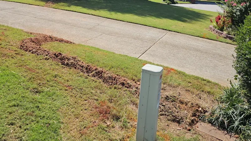 Trench dug along grassy slope near driveway, surrounding a white utility box in a residential neighborhood.