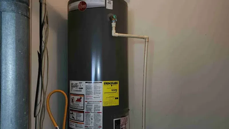 Gray Rheem water heater installed in a basement corner with pipes and an expansion tank above it.