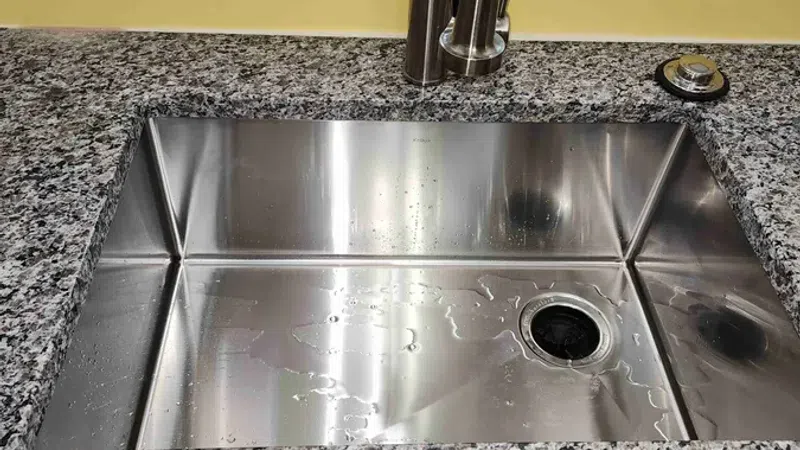 Stainless steel kitchen sink with water droplets, granite countertop, modern faucet, and yellow wall background.