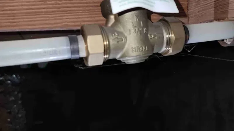 Close-up of a brass water valve with connecting pipes installed under a wooden beam in a dimly lit space.