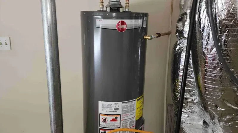 Gray Rheem water heater installed in basement with connected copper pipes and metal venting duct.