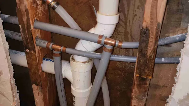 Exposed plumbing with PVC and PEX pipes inside a wall cavity, showing pipe joints and wooden studs.