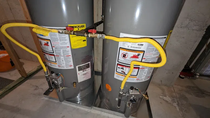 Two gray water heaters connected by yellow flexible gas lines in a concrete basement with visible warning labels.