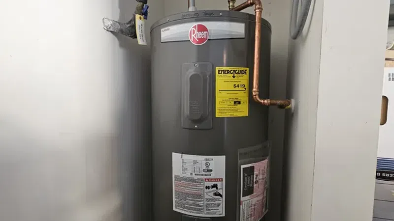 Gray Rheem water heater with copper pipes and energy guide label installed in a utility closet.