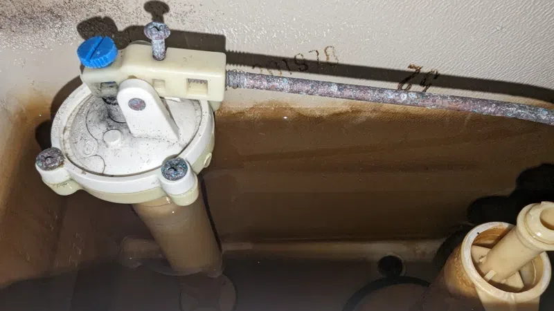 Close-up of a toilet tank interior showing the fill valve, water level, and plastic components with some rust and grime buildup.