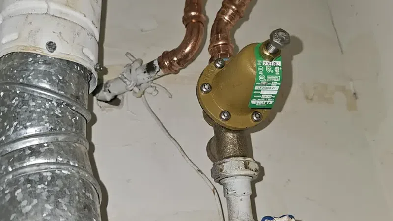 Copper and metal pipes with a valve and orange water shut-off tag in a utility room corner.