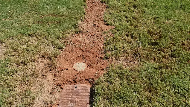 Narrow dirt trench cutting through a grassy lawn leading to a rectangular utility cover and circular marker.
