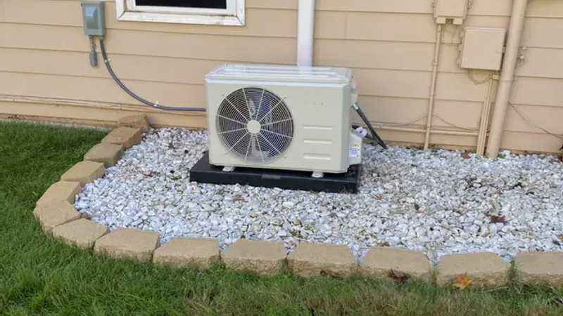 Outdoor air conditioning unit installed on gravel bed next to beige house siding and window.