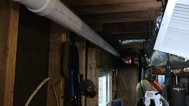 Basement workspace with exposed wooden beams, white PVC pipe, ductwork, and various tools and items on shelves