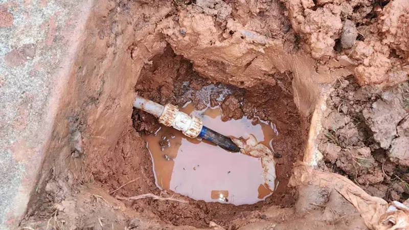 Open ground pit showing a leaking water pipe joint with muddy water pooling around it.