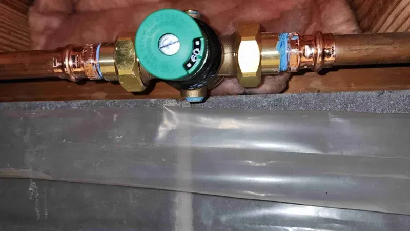Copper plumbing pipes connected to a green valve in an insulated ceiling with wooden beams and wiring.