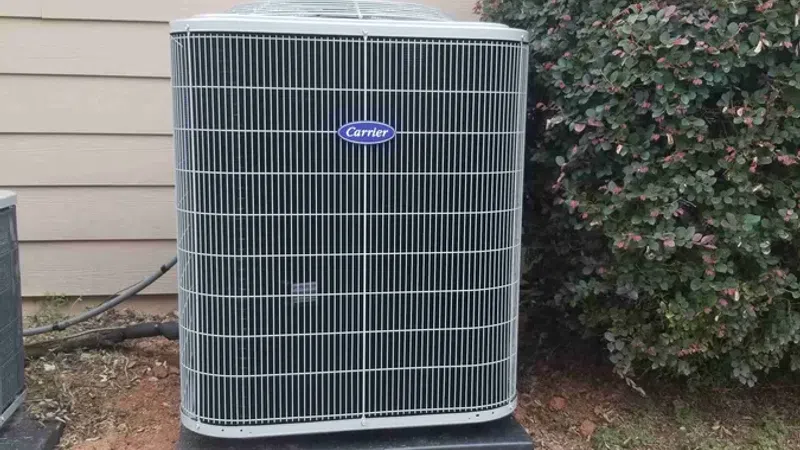 Carrier brand outdoor air conditioning unit installed beside a house and next to a bush on a grass lawn.