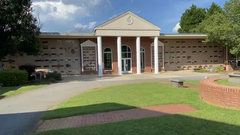 Brick building with white columns under a blue sky with large white clouds, surrounded by green trees and grass.