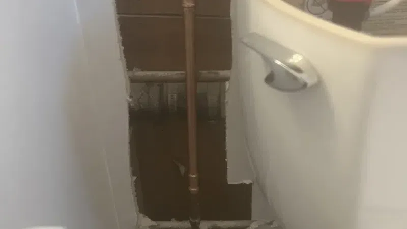 Exposed copper pipe behind wall near toilet with drywall cut out showing plumbing repair or inspection.