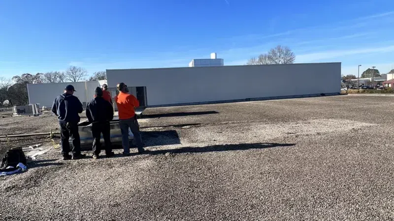 Three workers on a gravel rooftop watch an air conditioning unit being lifted by a crane on a clear sunny day.
