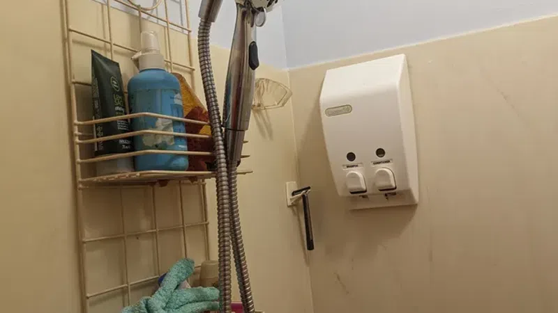 Bathroom shower area with a handheld showerhead, toiletries rack, soap dispenser, and cleaning tools on beige walls.