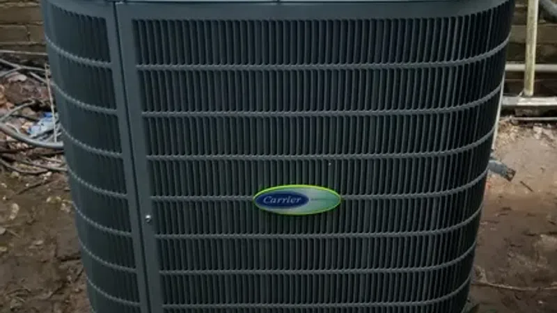 Outdoor Carrier air conditioning unit installed on a concrete slab beside a brick wall with surrounding dirt and plants.