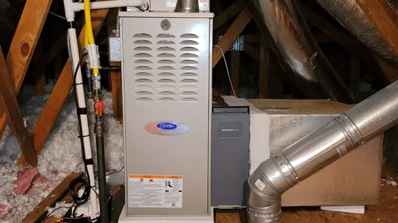 Residential heating furnace with ductwork and insulated pipes installed in an attic space with wooden beams.