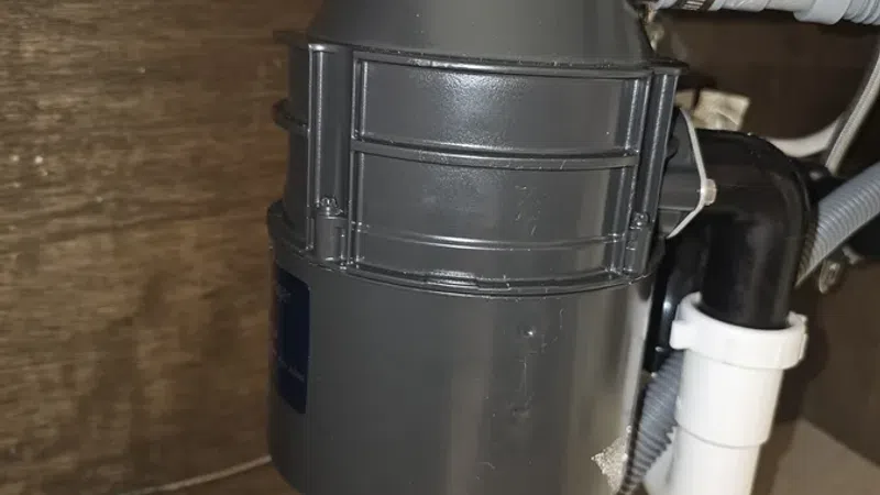 Close-up of a black kitchen sink garbage disposal unit installed under a wooden sink cabinet with connected plumbing.