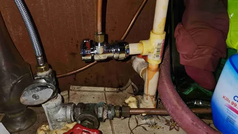 Close-up of household plumbing pipes, red valve, and cleaning gloves on floral patterned floor tiles under a sink.