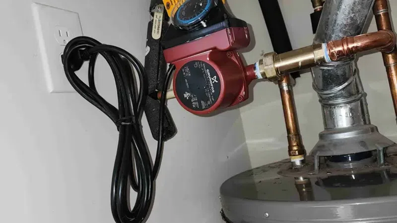 Red circulation pump attached to copper pipes on a water heater with insulation and electrical outlet nearby.