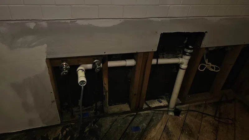 Exposed plumbing pipes and wooden studs behind partially removed drywall in a renovation site with damaged wooden flooring.