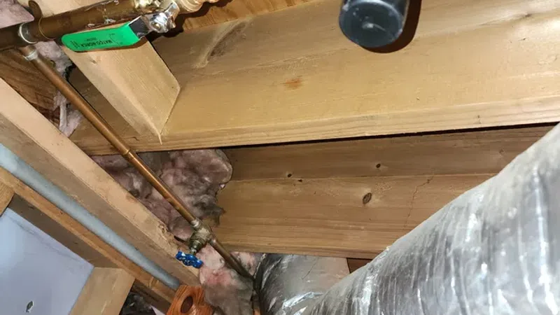 Copper plumbing pipes and insulated ductwork installed in a wooden attic with pink insulation material.