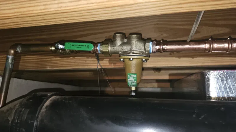 Brass water filter attached to copper pipes under a wooden floor with a black foam insulated pipe below.