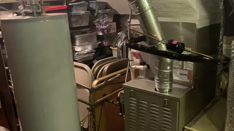 Utility room with water heater, furnace, ductwork, and air purifier unit surrounded by storage items and folding chairs.