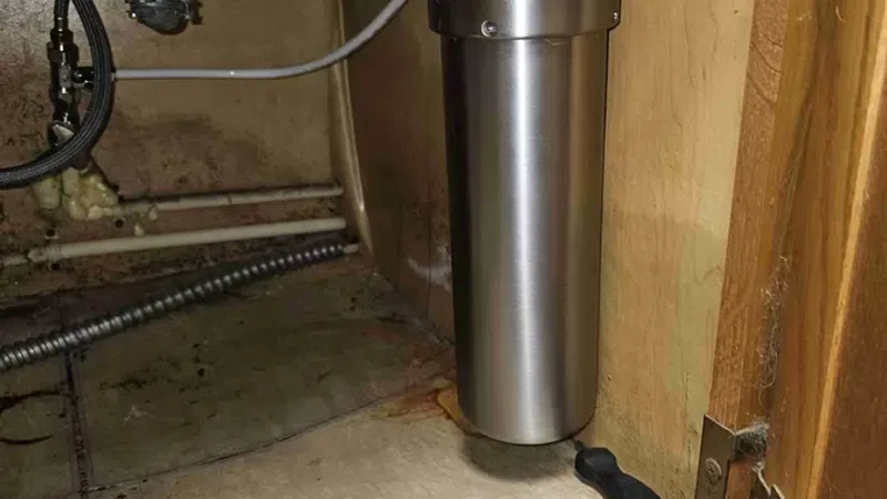 Under-sink view of a stainless steel water filter attached to plumbing in a wooden cabinet.