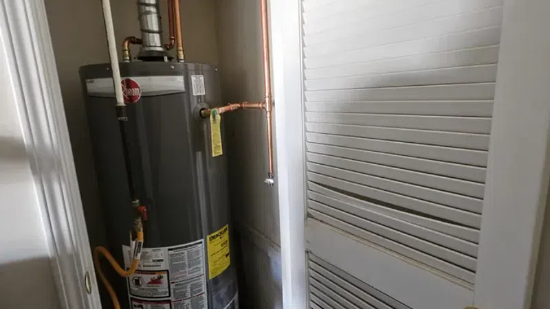 Tall gray water heater installed in a small utility closet with copper pipes and a white louvered door partially open.