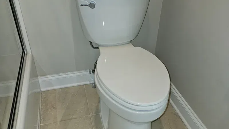 White porcelain toilet with closed lid in a beige tiled bathroom corner next to glass shower door.