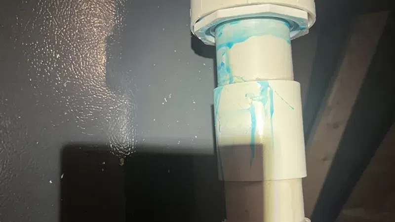 Close-up of white PVC plumbing pipes with blue primer at joints in a dimly lit basement wall corner.