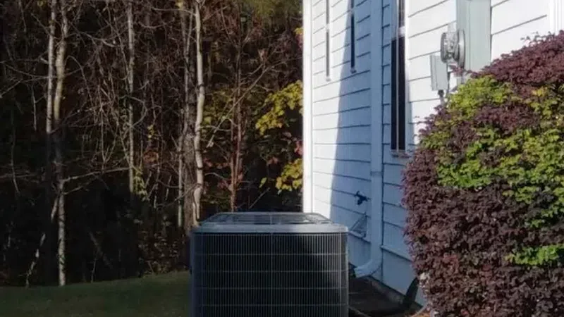 Outdoor air conditioning unit next to a white house with a green trash bin and white picket fence in fall.