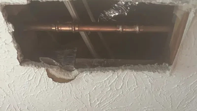 Rectangular hole in textured ceiling reveals copper pipes and foil ducting above the ceiling.