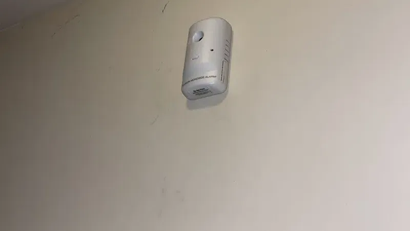 White smoke detector mounted high on a beige wall near a door frame in an interior room.