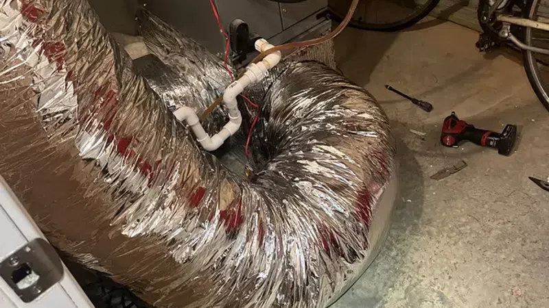 Flexible insulated duct connected to HVAC unit in basement with bike and tools nearby on concrete floor.