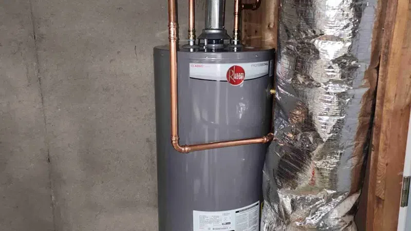 Gray Rheem water heater installed with copper pipes and insulation in a basement corner with concrete walls.