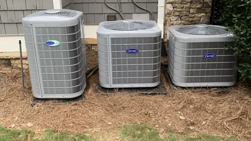 Three Carrier brand outdoor air conditioning units installed on ground with house siding and stone wall background.